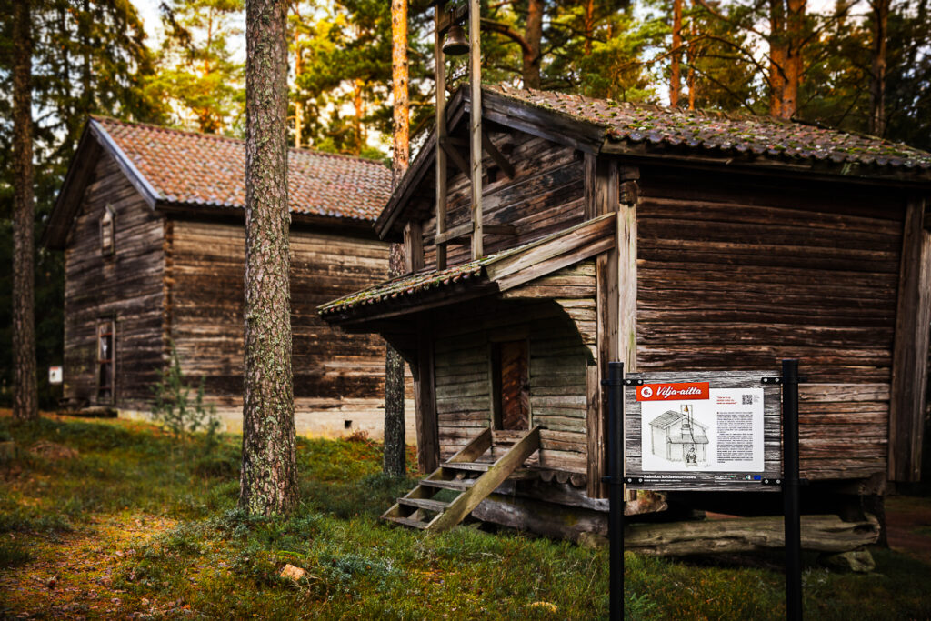 Kaksi museorakennusta metsäisessä maastossa. Toisen edessä on opastuskyltti, jonka otsikko Vilja-aitta