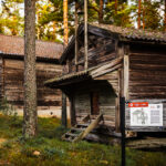 Kaksi museorakennusta metsäisessä maastossa. Toisen edessä on opastuskyltti, jonka otsikko Vilja-aitta
