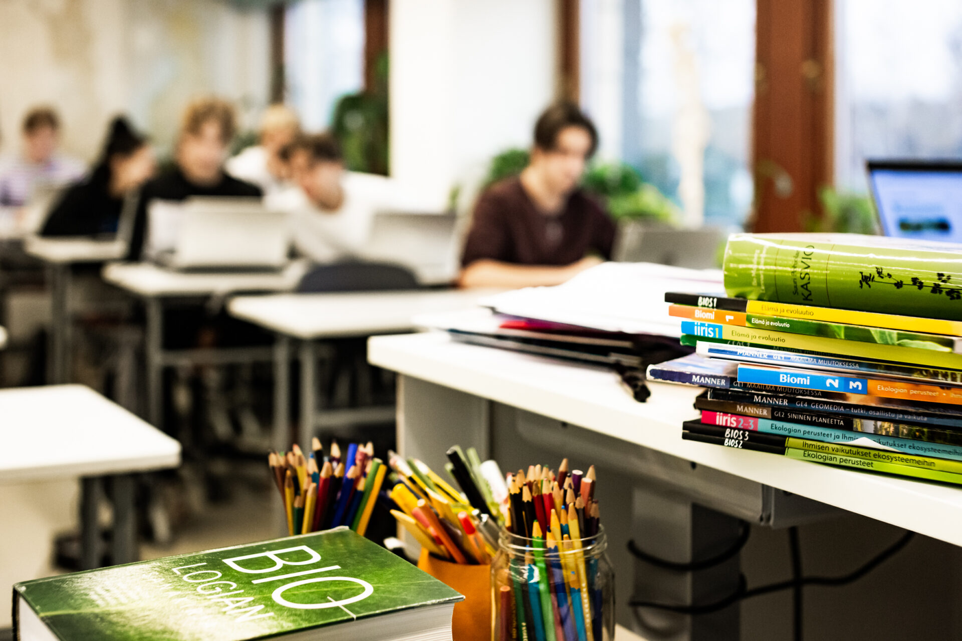 Etualalla työpöytä, jonka päällä on biologian oppikirjoja, taustalla näkyyn oppilaita sumennuttuna.