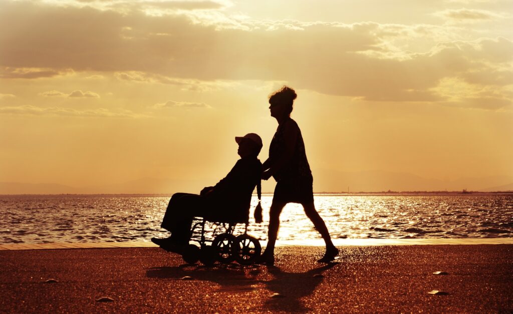 Nainen työntää pyörätuolissa olevaa vanhempaa naista meren rannalla.