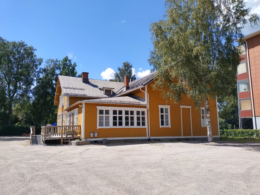Aurinkoisella säällä otettu kuva keltaiseksi maalatusta rakennuksesta, joka koostuu kolmesta osasta. Etualalla hiekkapohjainen pysäköintialue.
