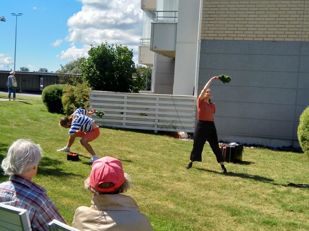 Tanssitaiteijat esiintymässä kauniina kesäpäivänä kerrostalon piha-alueella.