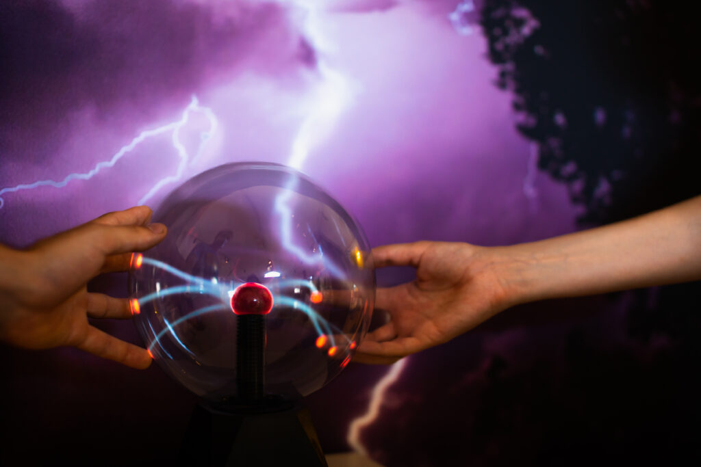 Kaksi kättä koskettaa plasmapallon pintaa. Taustalla näkyy sähköpurkaus.