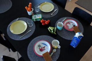 Lapsille katettu ruokapöytä, jossa lautasia, mukeja ja hieman vihanneksia ja juureksia.