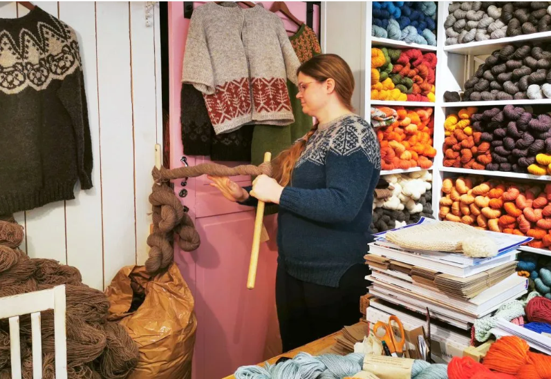 Nainen myymälässä käsittelemässä lankakieppiä. Taustalla näkyy myös villapaitoja ja runsaasti erivärisiä lankakieppejä. 