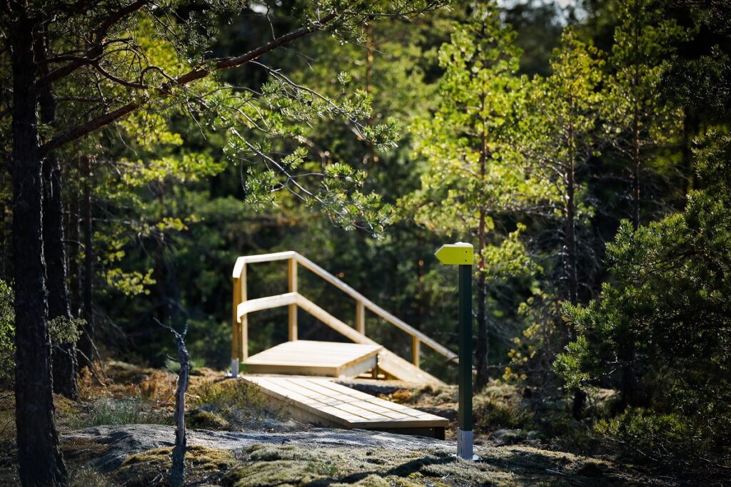 Metsäiseen ja rinne maastoon rakennetut portaat, jossa tukikaide. Etualalla kallioon kiinnitetty keltainen viittamerkki.