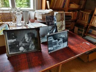 Pöydällä kehystettyjä valokuvia ja lasiesineitä. Taustalla kirjahyllyjä, joissa runsaasti kirjoja.