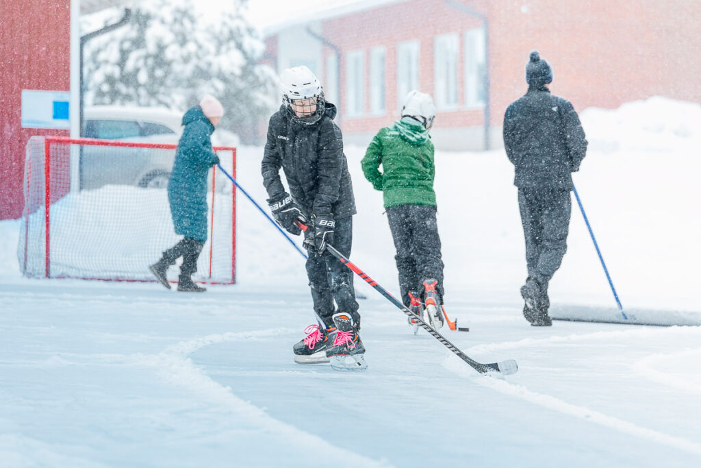 Luistelijoita ulkojääkentällä sekä lumisessa säässä jäätä kolalla puhdistava mies.