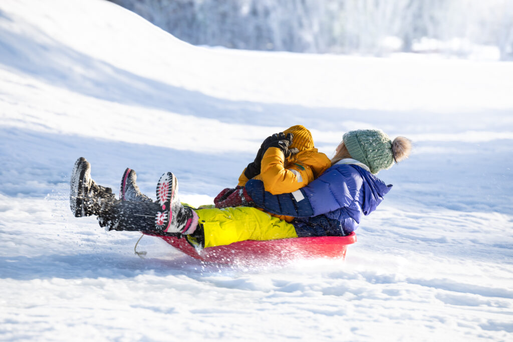 Punaisessa pulkassa kaksi ihmistä laskemassa mäkeä lumisessa maisemassa.