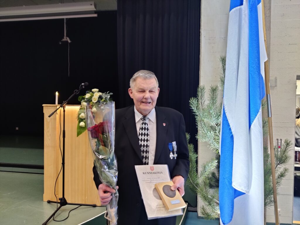 Mies ruusujen ja Paimio-mitalin kanssa, taustalla Suomen lippu.