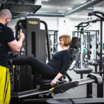 Kuntosalin työntekijäö neuvoo asiakasta kuntosalilaitteen käytössä.