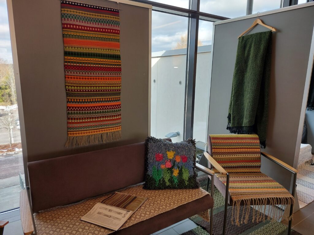 Erilaisia tekstiilitöitä näyttelyseinäkkeisiin ripustettuna että huonekalujen päällä.