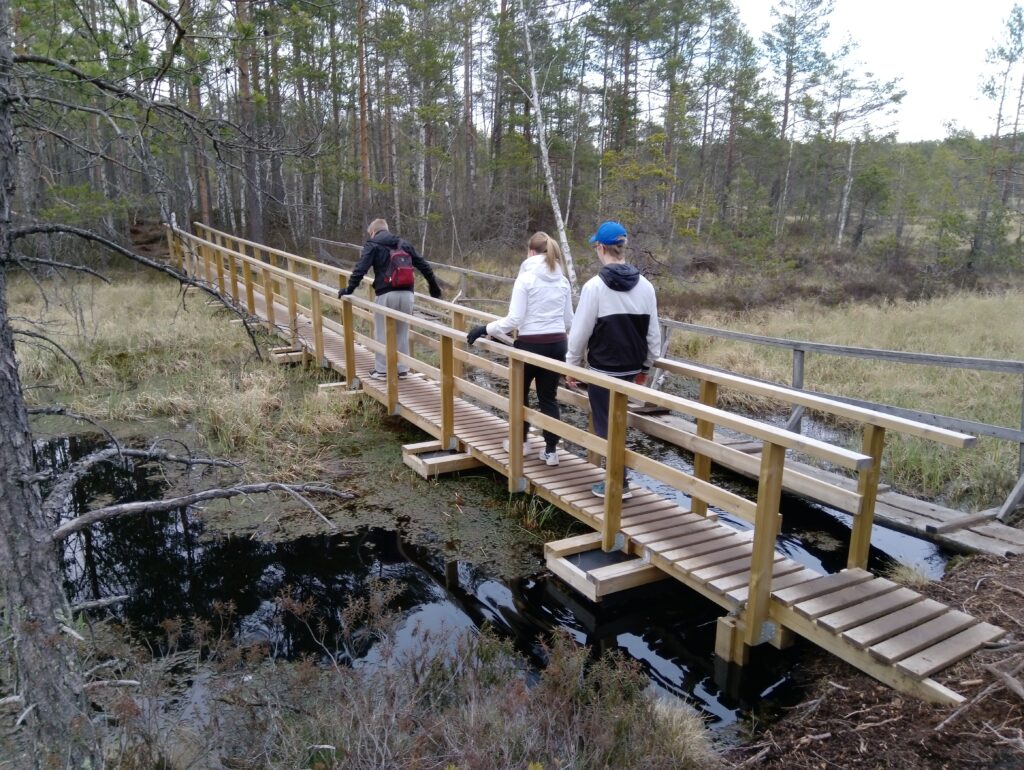Kolme ihmistä sillalla, joka kulkee suomaisen vesistön yli.