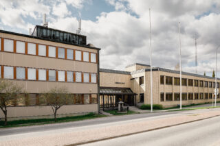 Ulkokuva rakennuksesta, jonka keskiosassa sisäänkäynti ja sen yläpuolella teksti Paimion lukio. Rakennuksen edustalla lipputankoja ja asvalttitie.