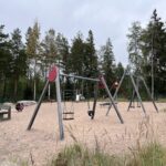 Hiekkapohjainen leikkipuisto välineineen metsä reunustalla.