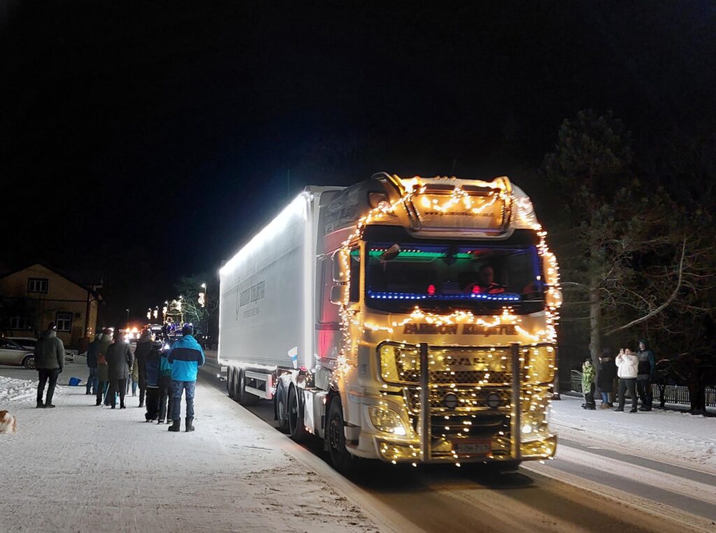 Iso kuorma-auto kulkee katua pitkin, jonka varrella ihmisiä. Auto on koristeltu erilaisin valoin.