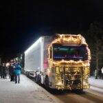 Iso kuorma-auto kulkee katua pitkin, jonka varrella ihmisiä. Auto on koristeltu erilaisin valoin.