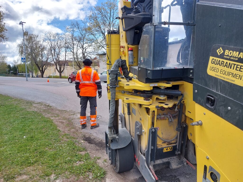 Kadun vieressä on keltainen korjausajoneuvo sekä oranssiin haalariin pukeutunut työntekijä. Taustalla olevalla tiellä kulkee autoja. Teien vieressä varoitusmerkki.
