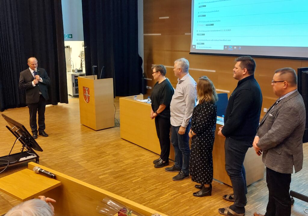 Kuusi henkilöä seisoo juhlasalin etuosassa. Vasemmalla seisovalla miehellä on kädessään mikrofoni.
