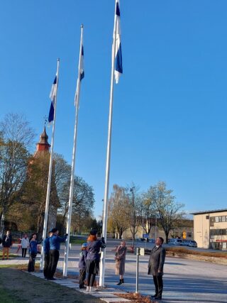 Kolme lipputankoon nostettua Suomen lippua kauniissa kevätsäässä. Lippuja nostamassa kuusi partiolaista ja heitä ohjaamassa kaksi miestä ja yksi nainen.