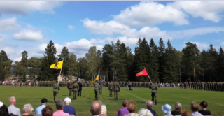 Kesäsäässä nurmikentällä runsaasti sotilaita. Lisäksi yksi keltapohjainen ja yksi punapohjainen lippu. Etualalla istuu runsaasti yleisöä.