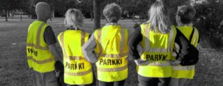 Viisi vapaaehtoista Parkki katupäivystäjää seisoo selkä kameraan päin päällään keltaiset liivit, jossa lukee selässä Parkki