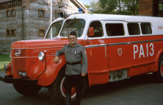 Puna-valkoinen vanha paloauto, jonka edessä seisoo mies palomiehen asusteissa.