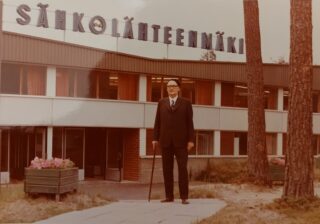 Baskeriin ja tummaan pukuun pukeutunut mies seisoo kävelykeppi toisessa kädessään seisoo rakennuksen edessä. Rakennuksen katolla lukee Sähkölähteenmäki.