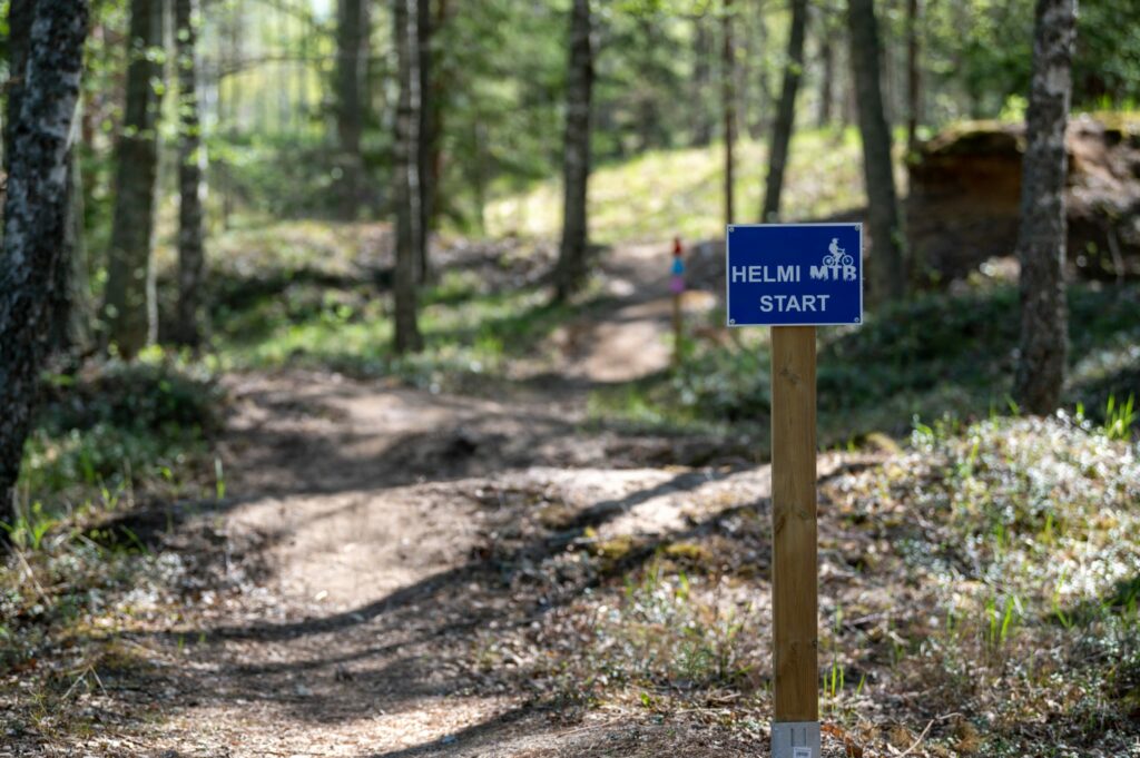 Helmi MTB Start kyltti ja metsäinen maisema, jossa menee maastopyöräily polku.