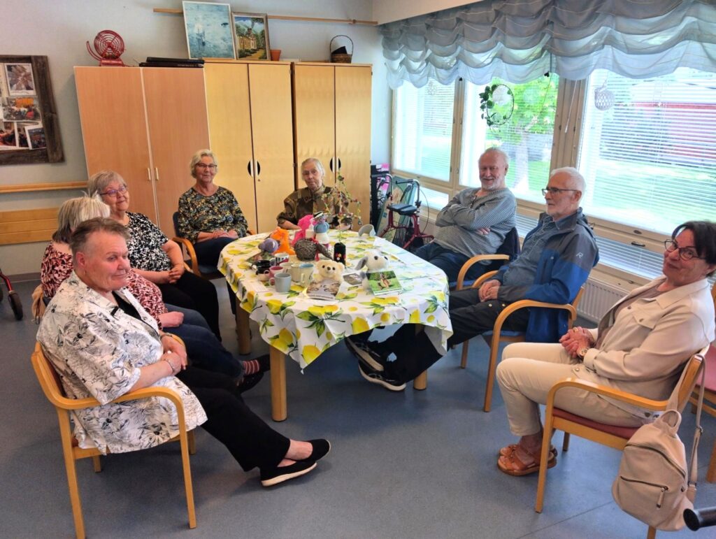 Ystäväpiiri ryhmä istuu pöydän ääressä, kuvassa taustalla kaapit ja ikkunat. Ryhmässä on kahdeksan ihmistä.