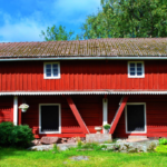 Kesäinen kuva kaksikerroksisesta, punaisesta luhtirakennuksesta, jonka lähettyvillä kasvaa myös lehtipuita.