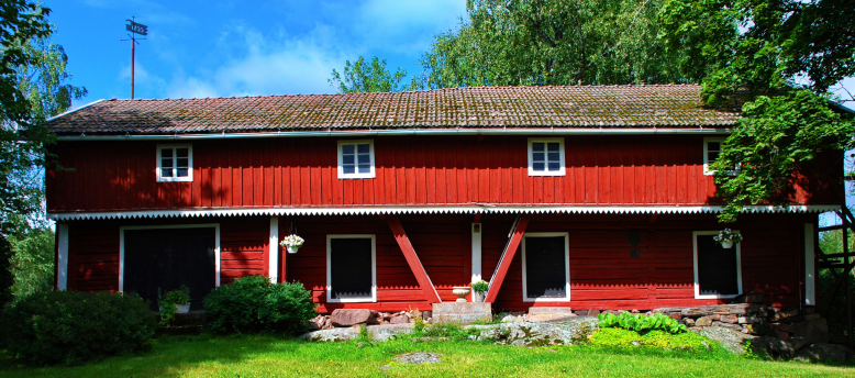 Kesäinen kuva kaksikerroksisesta, punaisesta luhtirakennuksesta, jonka lähettyvillä kasvaa myös lehtipuita.