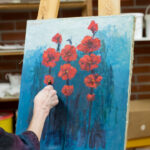 Maalarin käsi maalaa sinistä taulua jossa punaisia kukkia