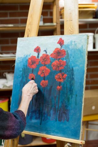 Maalarin käsi maalaa sinistä taulua jossa punaisia kukkia