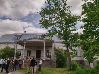 Kesäsäässä ihmisiä seisomassa ison rakennuksen pihamaalla.