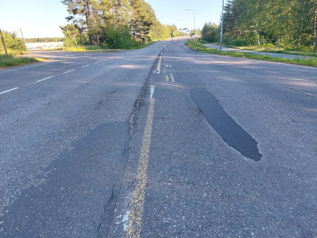 Etualalla osin huonokuntoista päällystettä, taustalla näkyy tieopasteita.