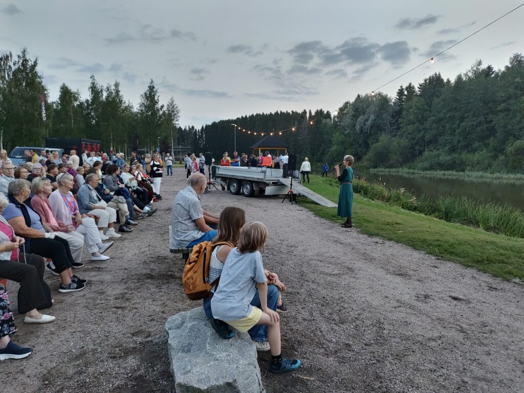 Ulkoilmatapahtuma joen tuntumassa. Yksi henkilö puhuu mikrofoniin ja runsaasti ihmisiä istuu penekeillä yleisönä.