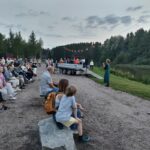 Ulkoilmatapahtuma joen tuntumassa. Yksi henkilö puhuu mikrofoniin ja runsaasti ihmisiä istuu penekeillä yleisönä.