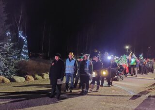 Pimeässä talvisäässä runsaasti ihmisiä ryhmityneet jonoon. Osalla on mukana lyhtyjä ja lippuja.