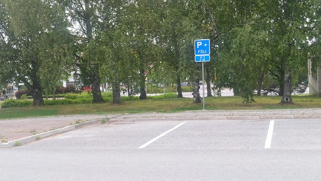 Asvaltoidun parkkialueen reunalla sinipohjainen liikennemerkki , jossa merkinnät P, +Föli, 12h ja 2 ap eli asiakaspaikkaa.