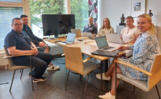 Kuusi henkilöä istumassa kokoushuoneessa pöydän ympärillä. Kaikkien edessä kannettavia tietokoneita.