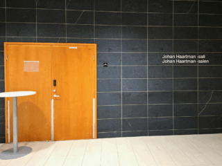 Tummapintainen seinä sekä vaalenruskeat oovet. Seinässä teksti Johan Haartman -sali, Johan Haartman -salen.