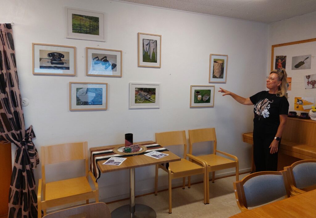 Naine esittelee huoneen seinille ripustettuja valokuvatauluja.
