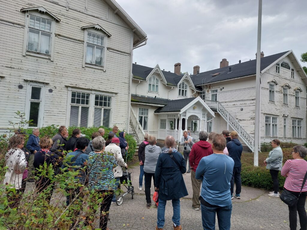 Valkoisen, useasta osasta koostuvan rakennuksen edessä paljon ihmisiä kuuntelemassa oppaan esitystä.