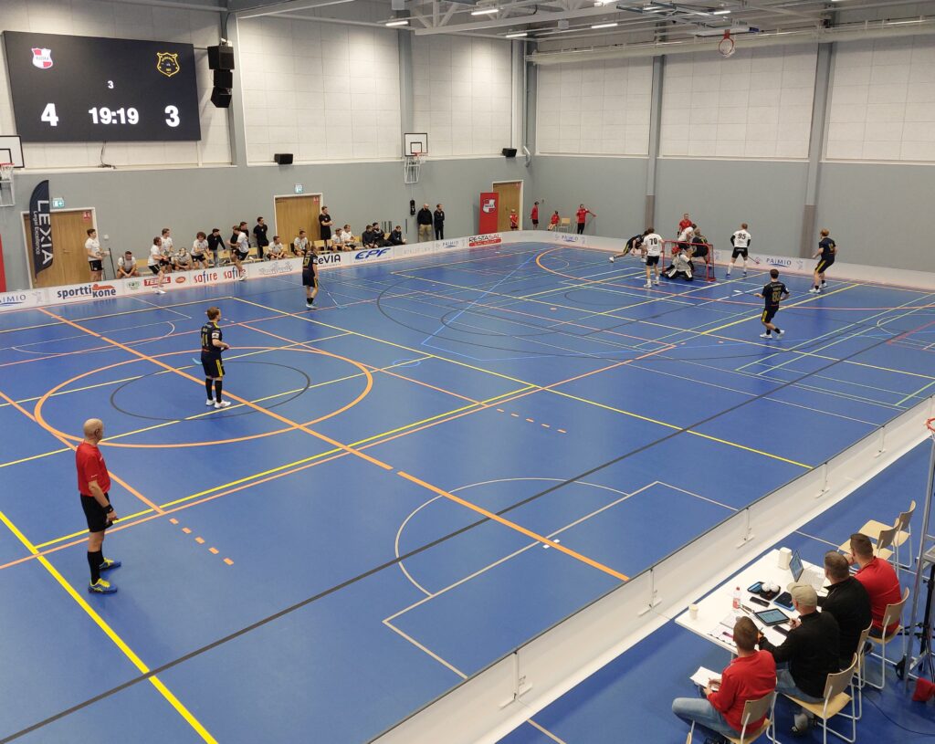Valkoisissa ja mustossa peliasuissa pelaaajia sisäliikuntahallissa, jossa sininen lattia. Seinällä näkyy pelikello sekä ottelutilanne 4-3.