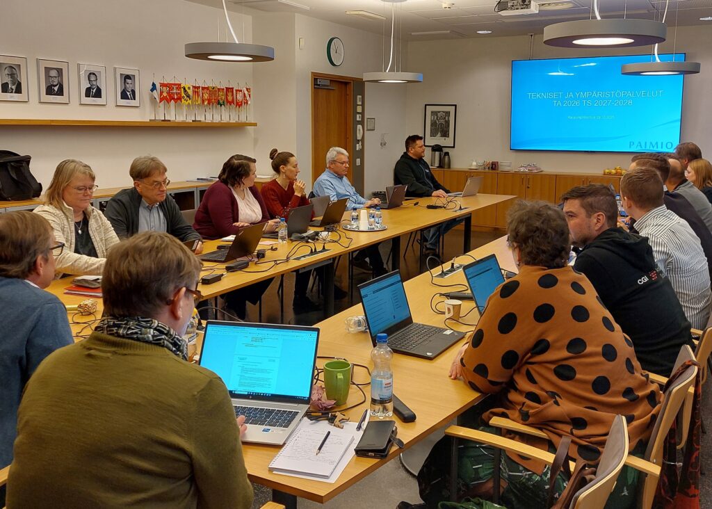 Runsaasti ihmisiä istumassa kokouspöydän ympärillä. Takaseinällä näkyy tv-näyttö.