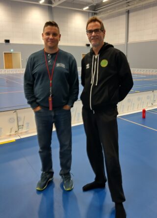 Kaksi miestä seisomassa siniseksi maalatun pelikentän reunalla.