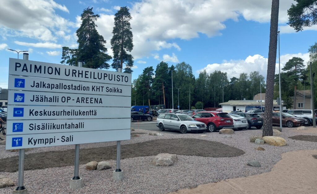Etualalla kyltti, jonka pääotsikkona on Paimion urheilupuisto ja sen alla keskeiset kohteet urheilupuiston alueella. Taustalla pysäköintipaikalla olevia autoja ja muutama rakennus.