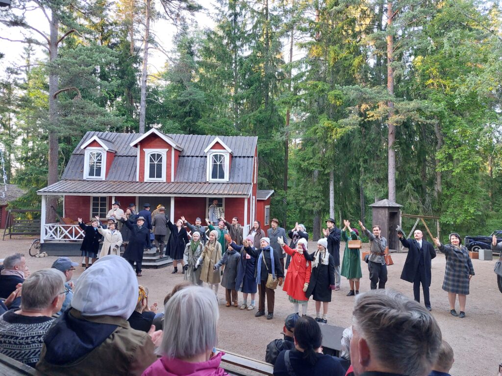 Punaisen rakennuksen edessä kesäteatterinäyttämöllä seisomassa runsaasti ihmisiä esiintymisasuissa. Etualalla näkyy katsomossa olevaa yleisöä 