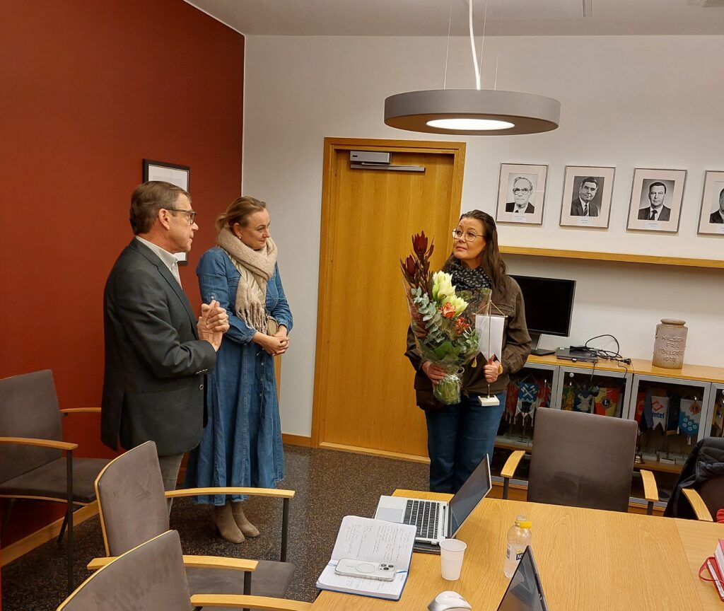 Kokoushuoneessa kolme henkilöä seisomassa. Oikealla olevalla on sylissään kukkia ja pöytäviiri.